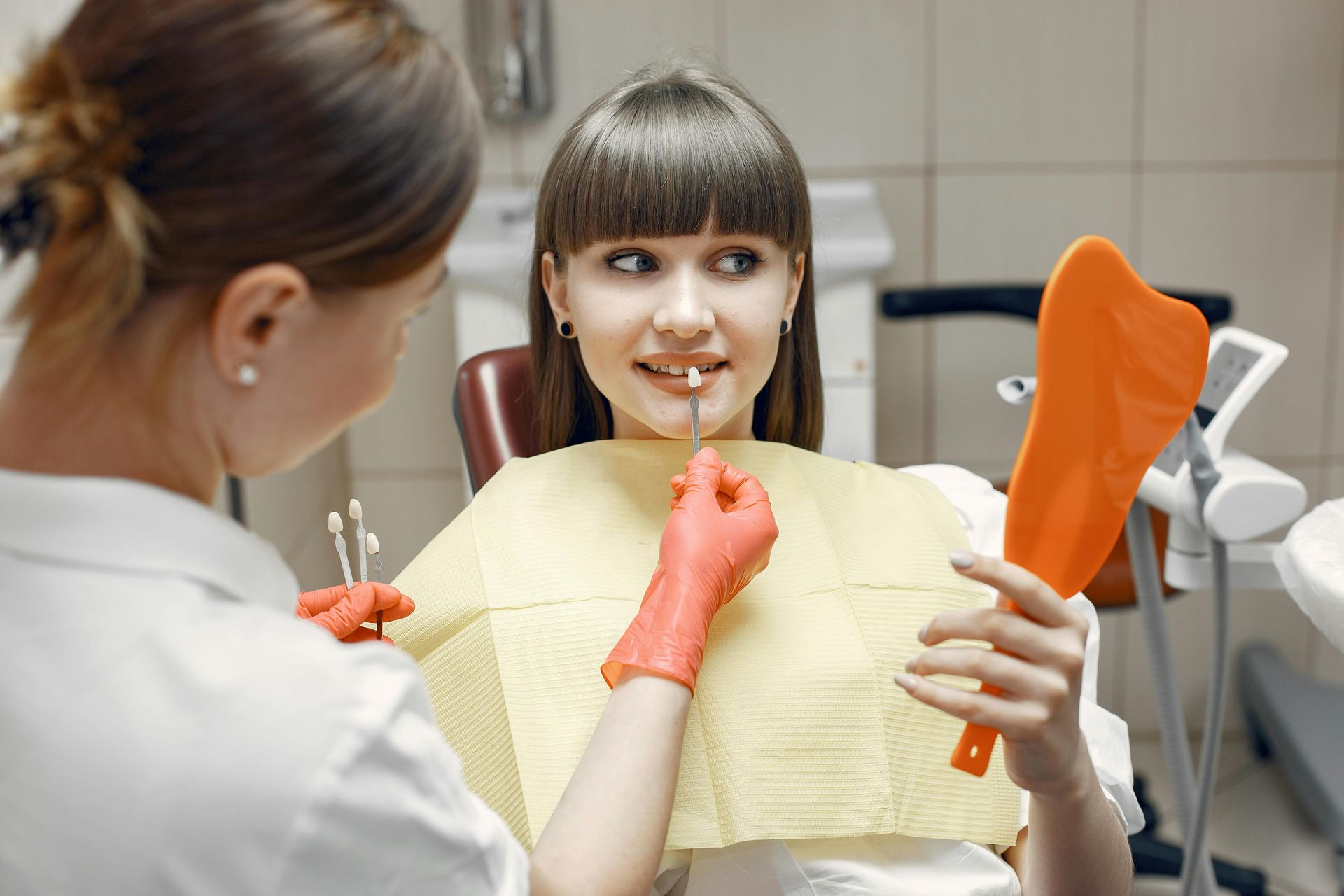 A dental professional holding a shade guide to a patient’s tooth while the patient holds a mirror in an exam room.