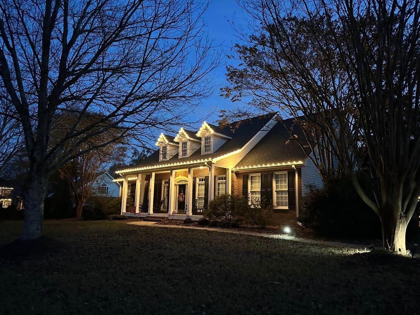 House at dusk with white Christmas lights on the roof and porch; trees frame the building, lawn in the foreground.