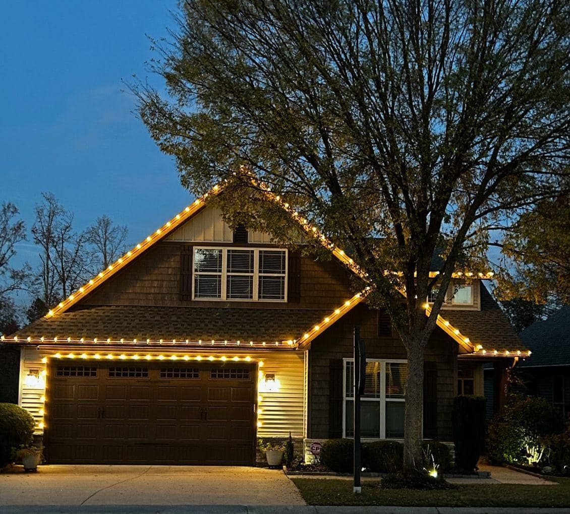 House decorated with yellow Christmas lights, brown roof, at dusk.