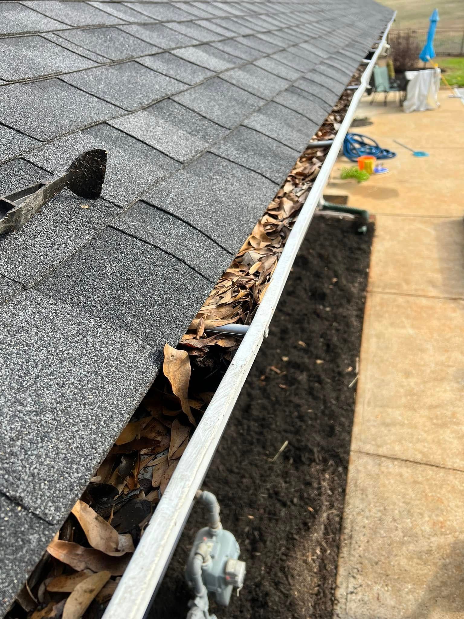 Gutter filled with leaves and debris, next to a dark mulch bed and a gray shingle roof.
