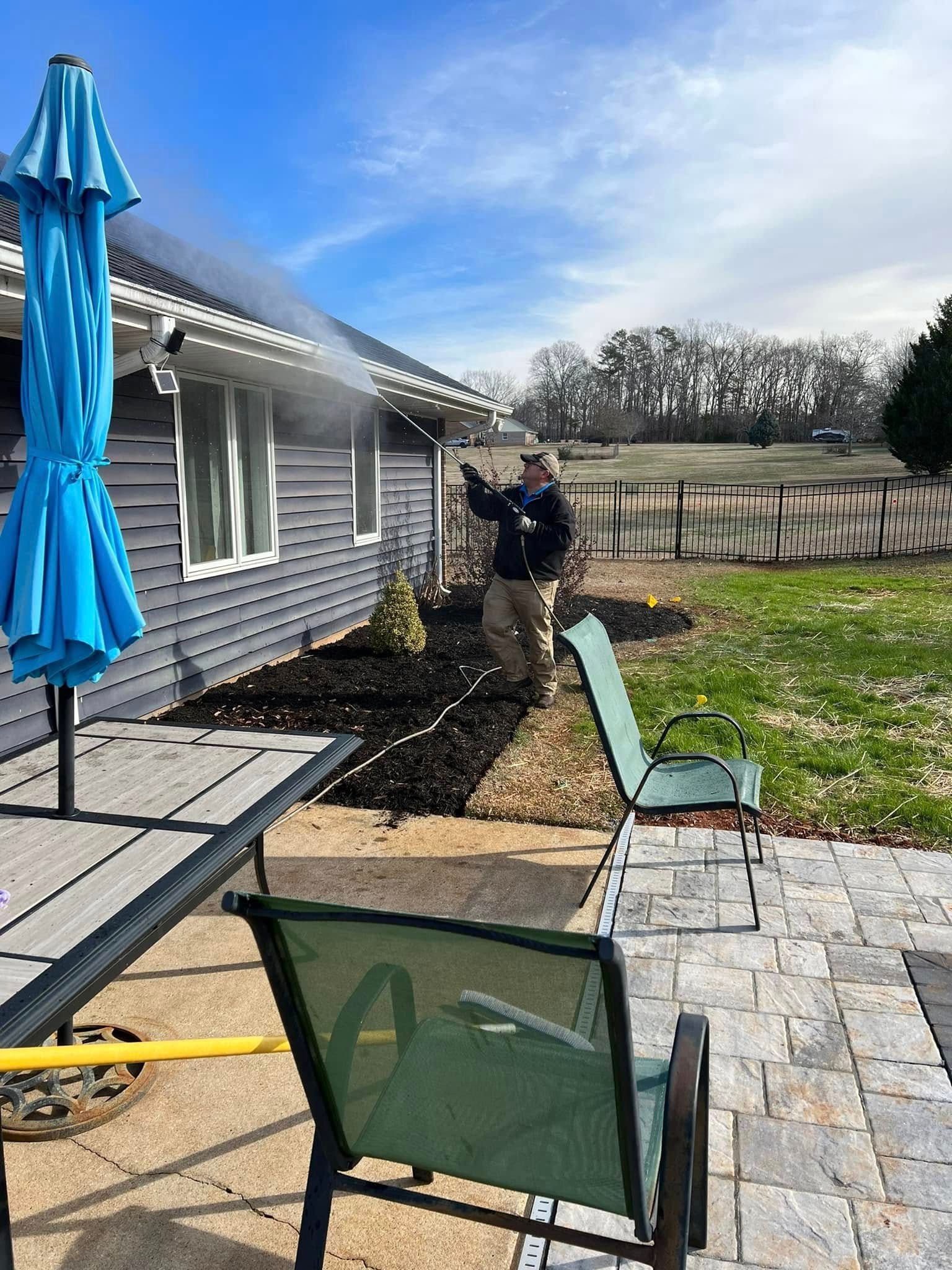 Man spraying a house with a pressure washer. Gray siding, blue umbrella, green chairs, sunny day.