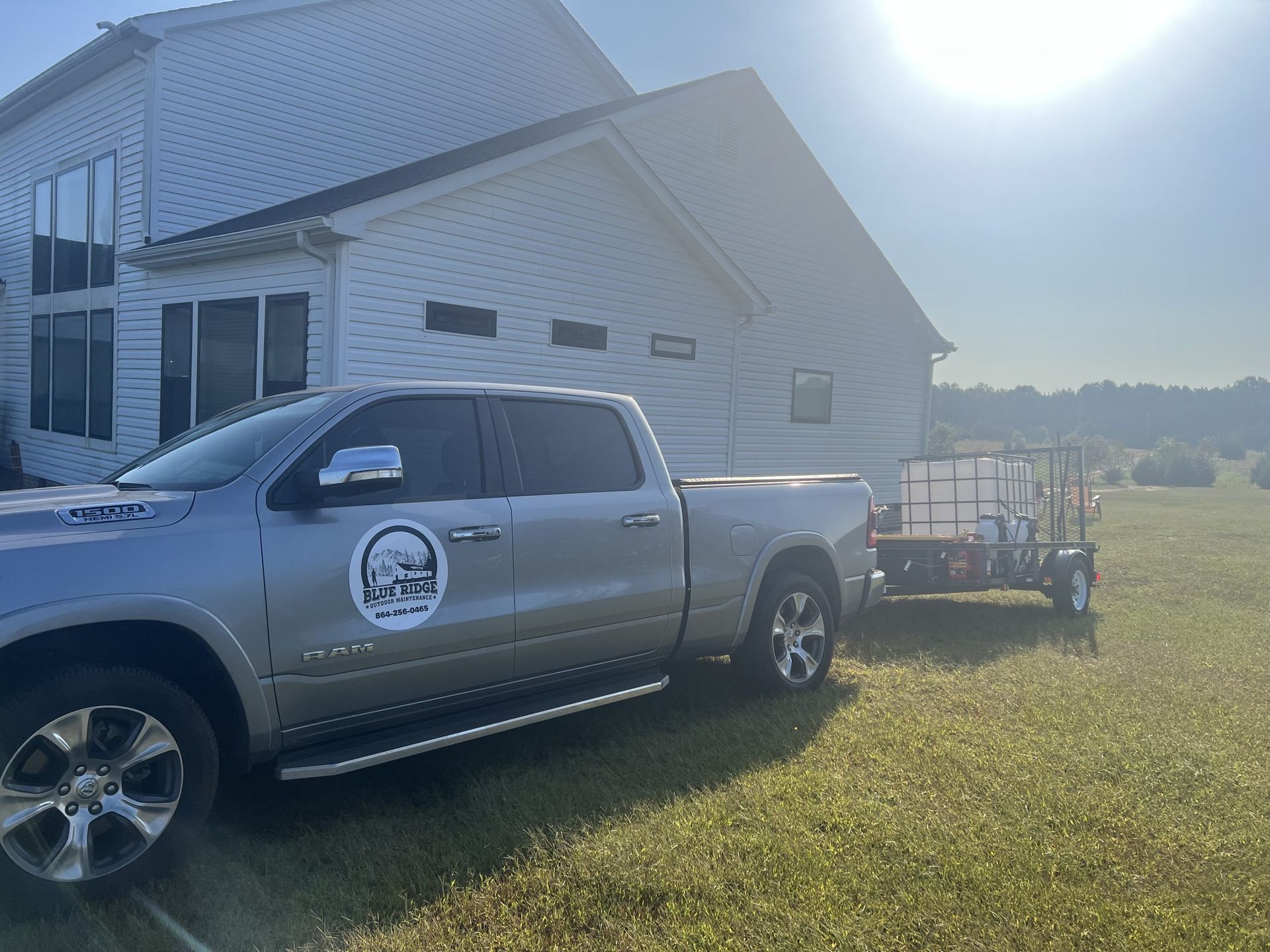 A silver pickup truck towing a trailer with a water tank parked on grass next to a house in daylight.