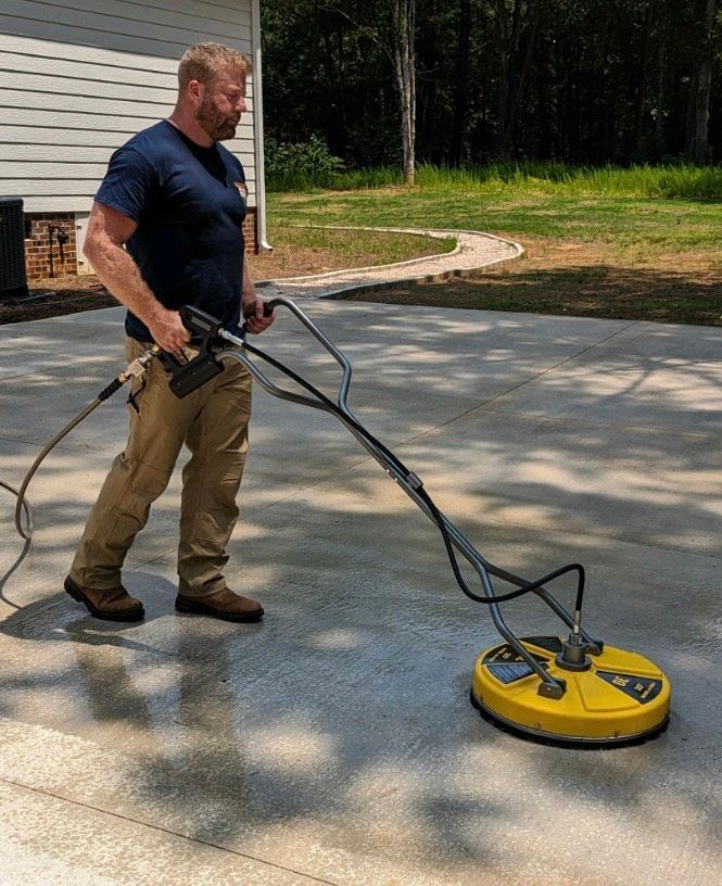 Man in a blue shirt using a yellow surface cleaner on a concrete driveway outdoors.