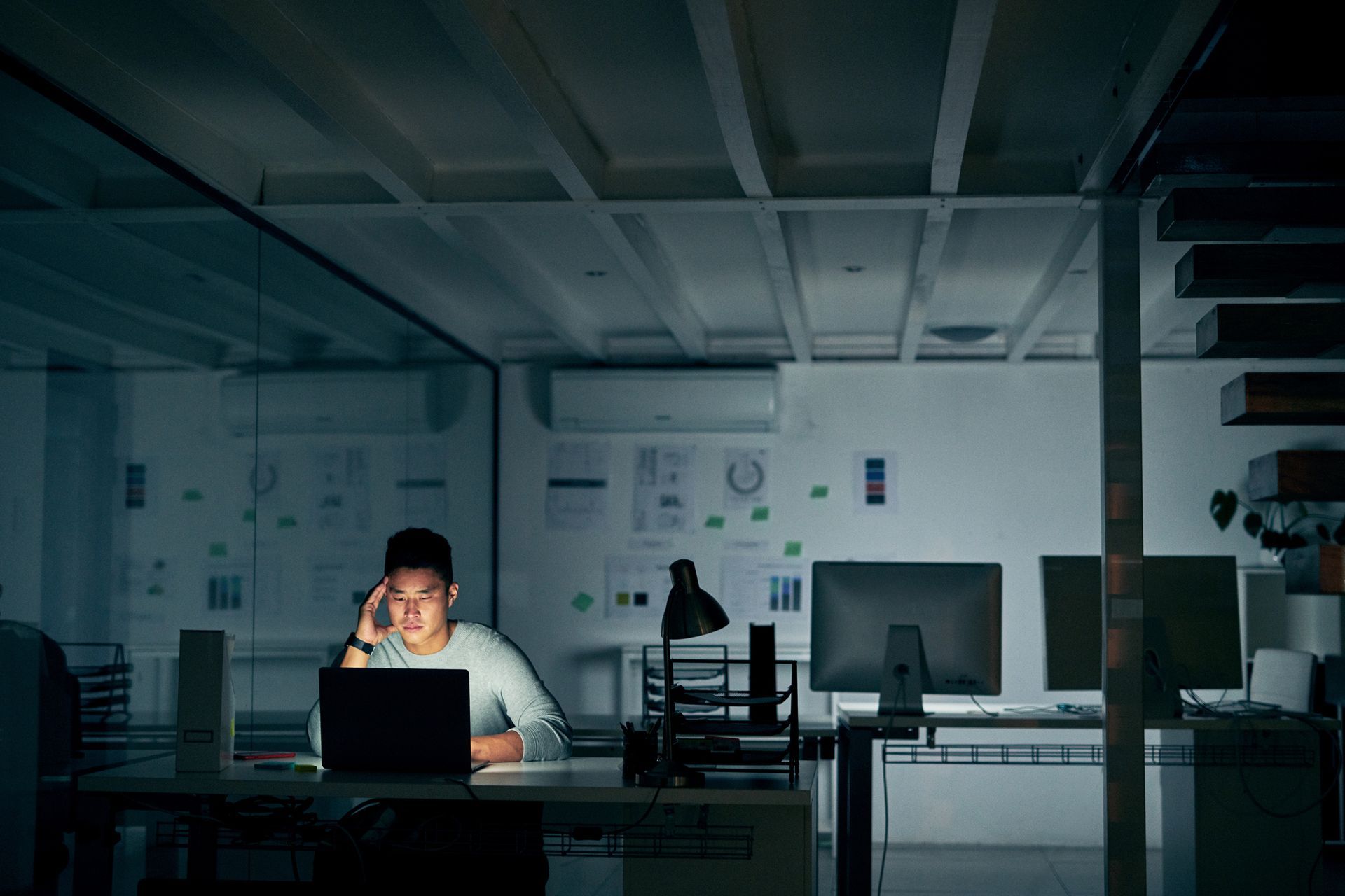 A man is sitting at a desk in an office with a laptop and talking on a cell phone.