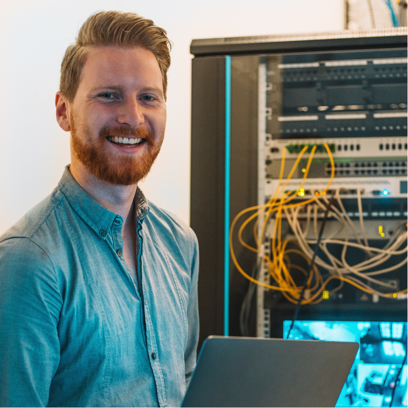 A man with a beard is holding a laptop in front of a server rack