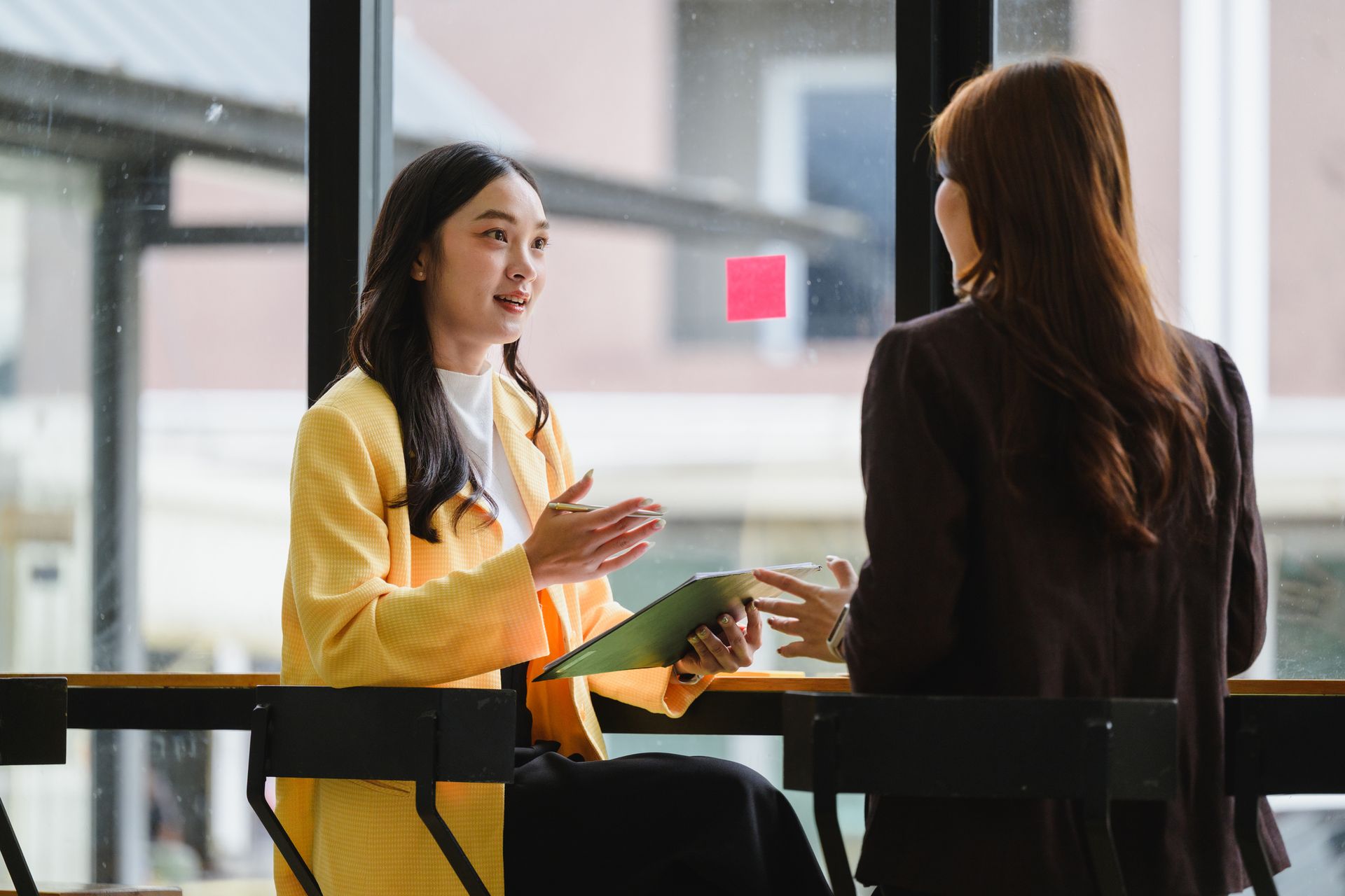 Two women talking at a table in a bright indoor café, one in a yellow jacket gesturing.