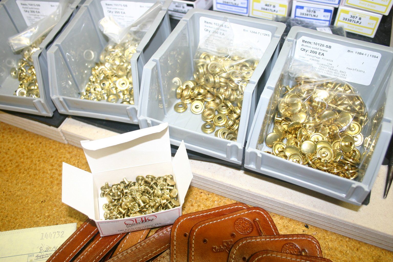 Bins of all brass hardware and a box of rivets, alongside leather ClubShields on a work surface.