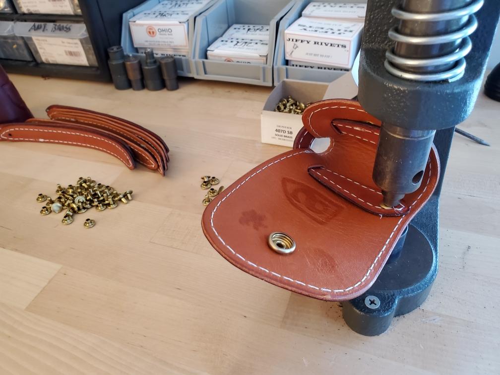 A leather item being riveted with a press machine in a workshop. Brass rivets and leather pieces are present.