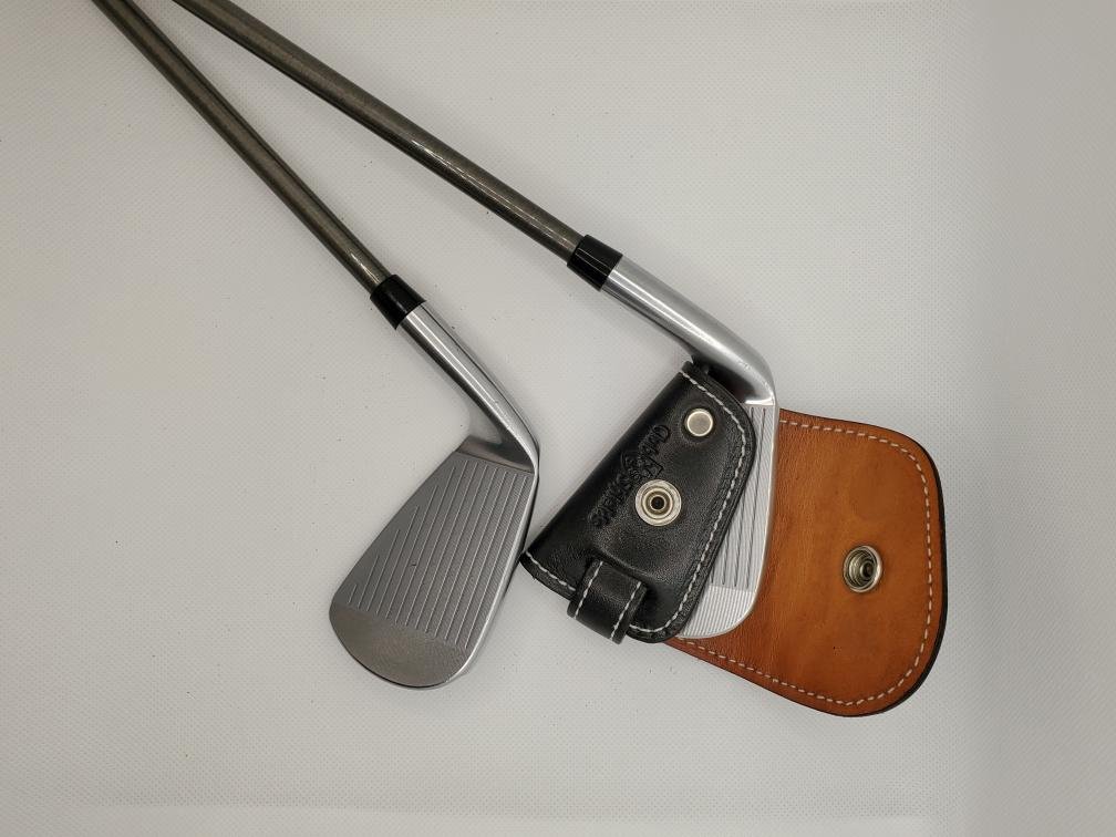 Two golf clubs next to a brown leather head cover on a white surface.