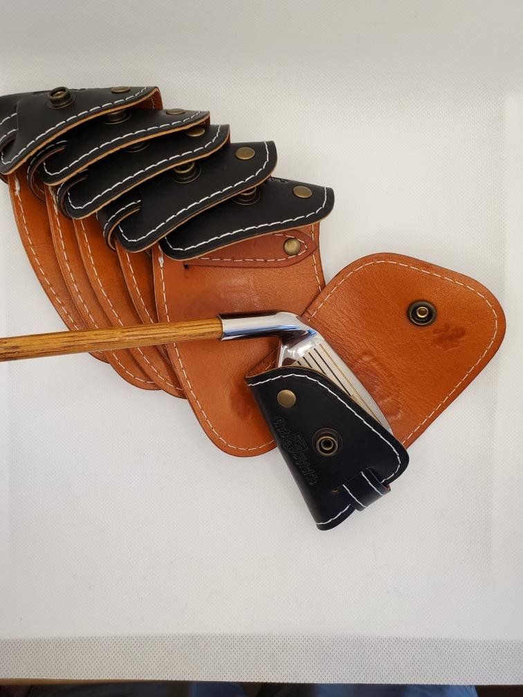 Golf club heads in leather covers, some stacked on brown leather pouch, on white surface.