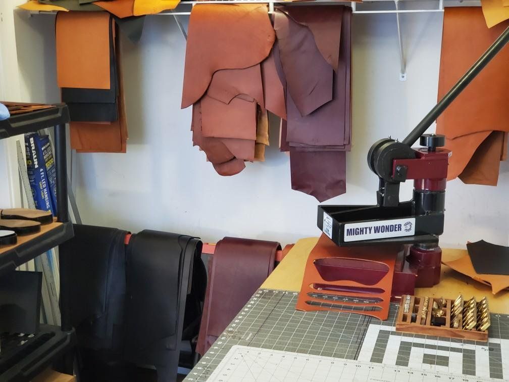 Leather workshop with press and materials: various leather colors hanging, cutting mat, and tools.