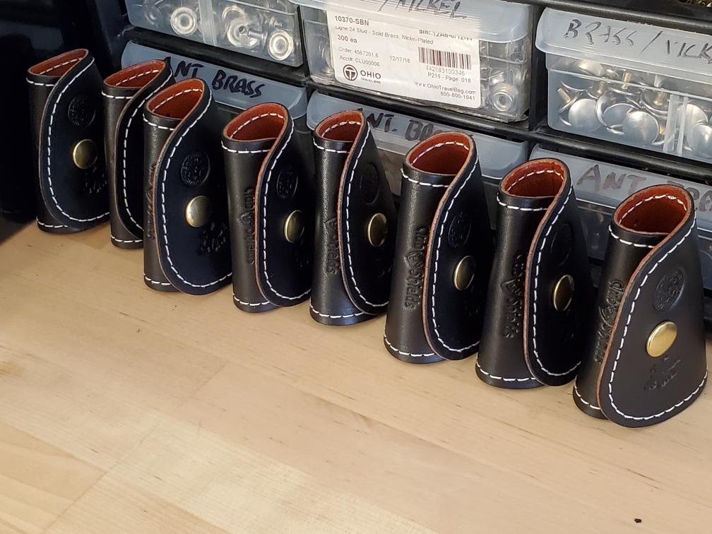 Nine black leather ClubShields covers in production with white stitching and brass buttons, on a wooden surface.