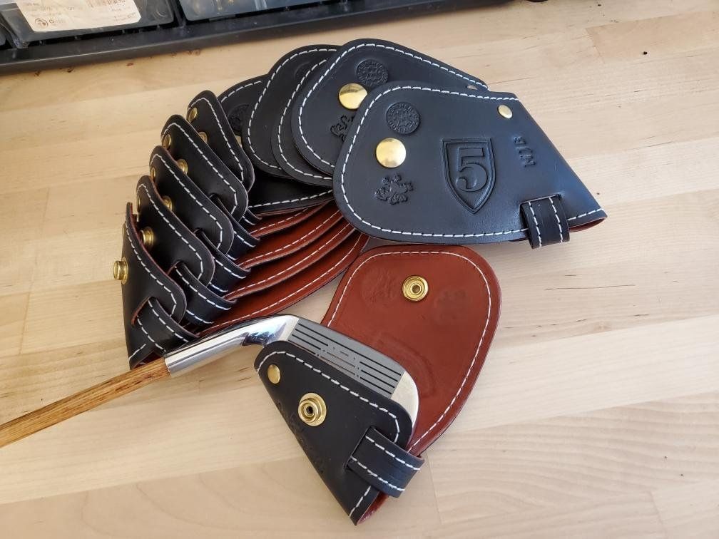A finished set of black leather golf club covers with white stitching and brass snaps, stacked with a golf club.