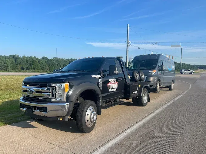 Black tow truck towing a gray van on a paved road under a blue sky.