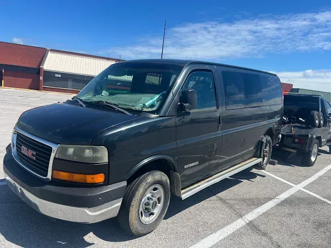 Black GMC Savana van parked on asphalt on a sunny day. A flatbed tow truck is behind it.