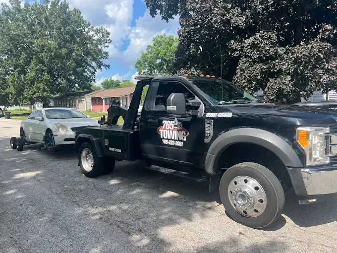Black tow truck towing a silver car on a paved road under a partly cloudy sky.