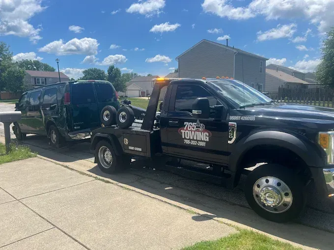 Black tow truck towing a dark green van on a sunny day in a residential neighborhood.
