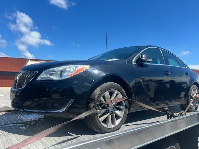 Black Buick Regal sedan strapped to a flatbed tow truck on a sunny day.