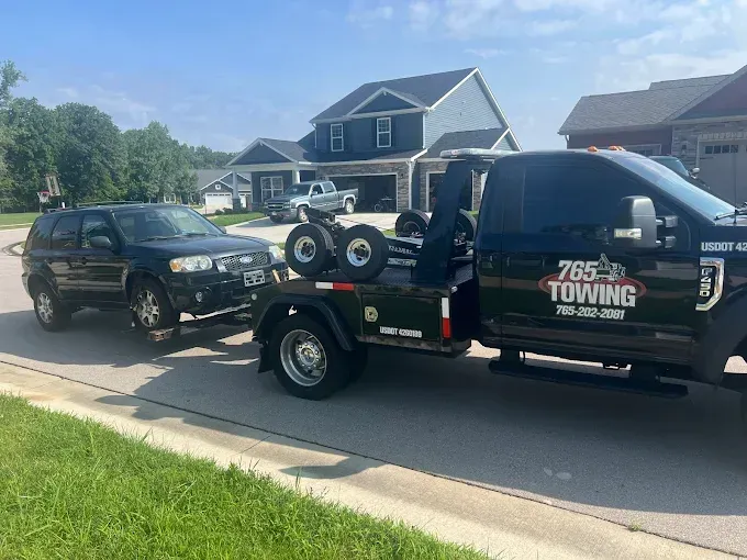 Black SUV being towed by a black tow truck on a residential street. Bright day, blue sky.