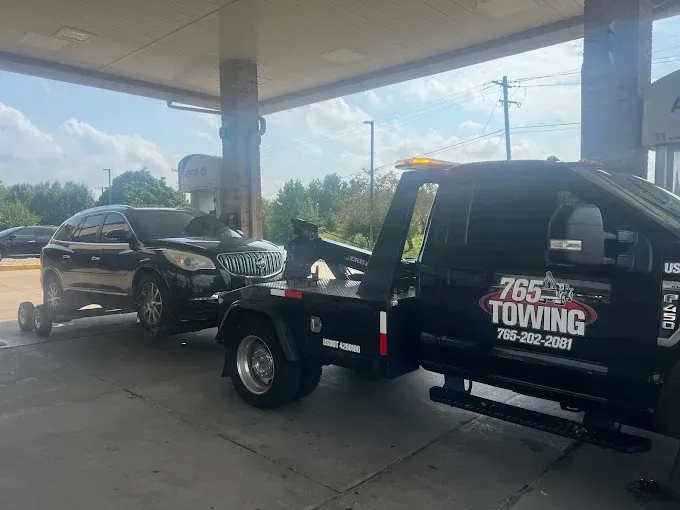 Black SUV being towed by a black tow truck at a gas station under a canopy.