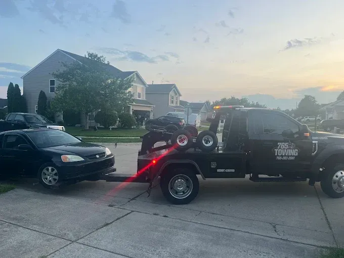 Tow truck pulling a black car with a small black ATV on the flatbed, parked in front of houses at dusk.