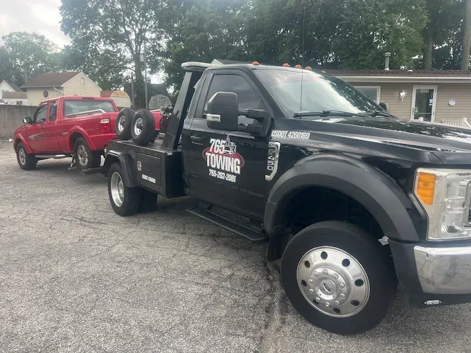 Black tow truck towing a red pickup truck on a paved lot. The truck has 