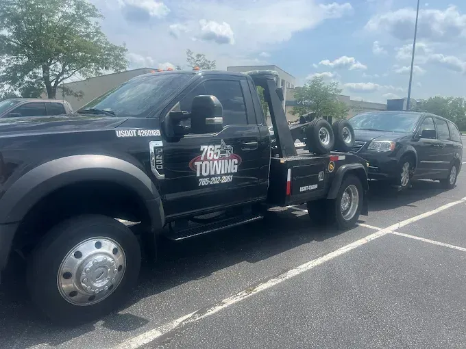Black tow truck towing a dark minivan on a sunny day in a parking lot. The truck has 