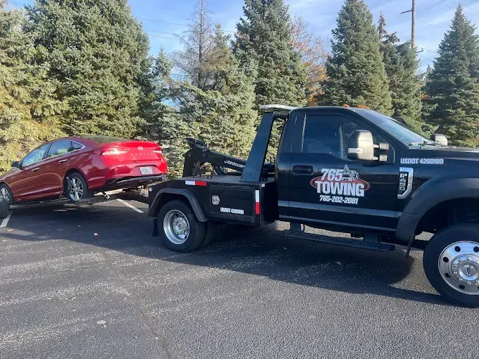 A red car is towed by a black tow truck in a parking lot on a sunny day.