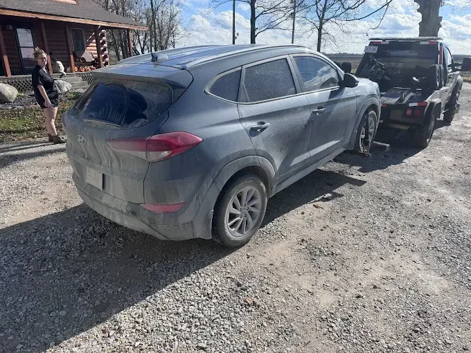 Gray SUV being towed by a tow truck on a gravel road, person standing near it.