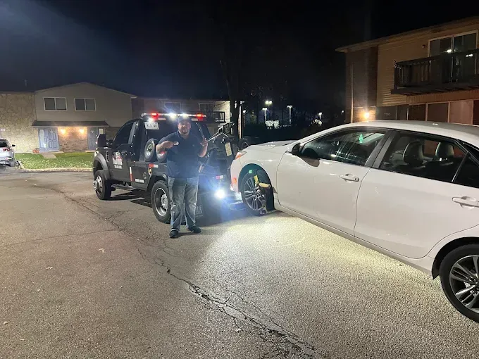 Tow truck towing a white car at night. Person stands near truck. Buildings in background.