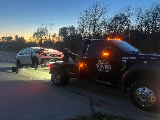 Tow truck towing a damaged white car on a road at dusk.
