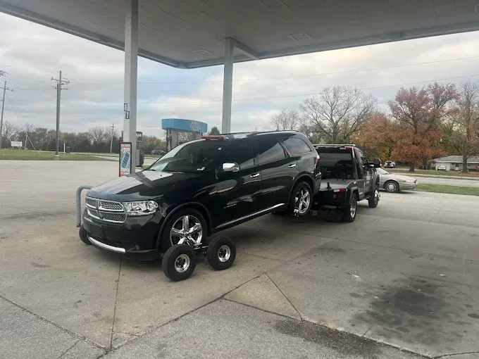 Black SUV being towed by a smaller black vehicle at a gas station, tires on tow wheels.