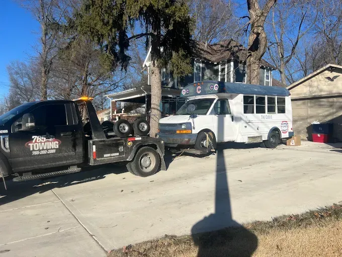 Tow truck towing a white bus on a concrete driveway in front of a house.