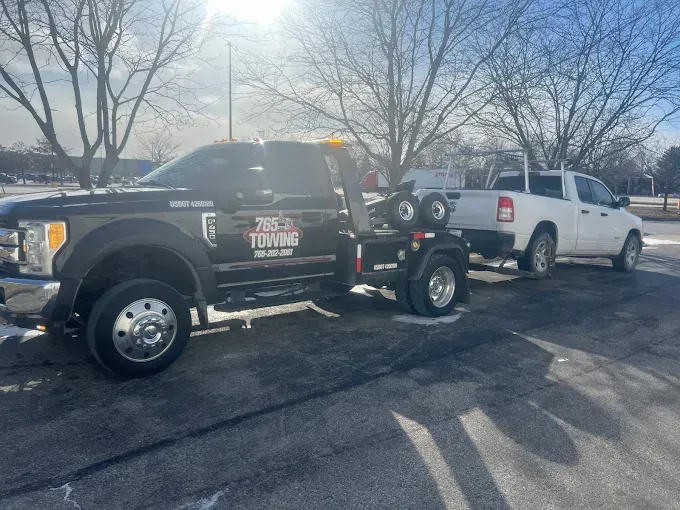 Black tow truck towing a white pickup truck on a paved lot in winter.