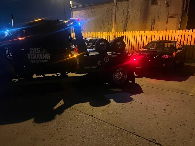 Tow truck towing a black car at night, lights flashing.
