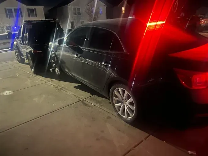 Black sedan being towed by a tow truck on a residential street at night, illuminated by flashing red lights.