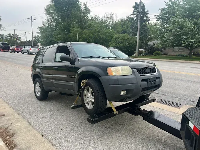 Black SUV being towed on a flatbed trailer on a street next to a sidewalk and parked cars.