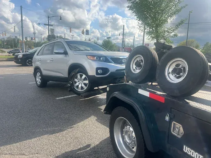 Silver SUV being towed by a tow truck on an asphalt road. Two spare tires are attached to the truck.