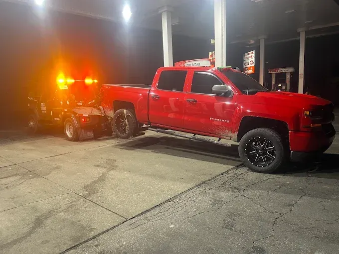 Red pickup truck being towed by a tow truck at a gas station, night.