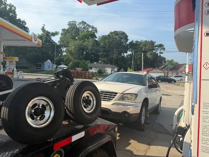 White car at a gas pump with a flatbed trailer hauling truck tires next to it at a gas station.