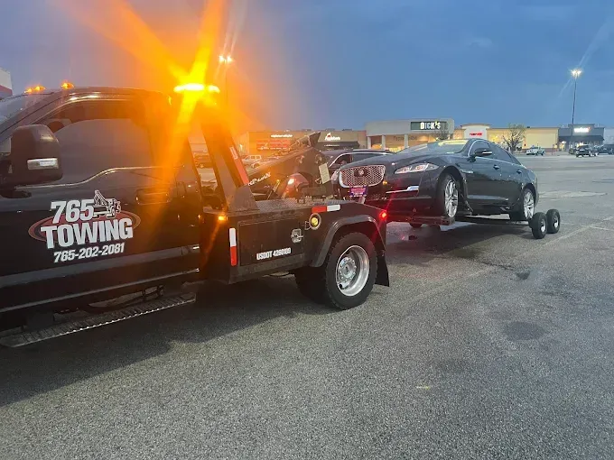 Tow truck towing a car in a parking lot. The truck has flashing amber lights, and the car is dark gray.