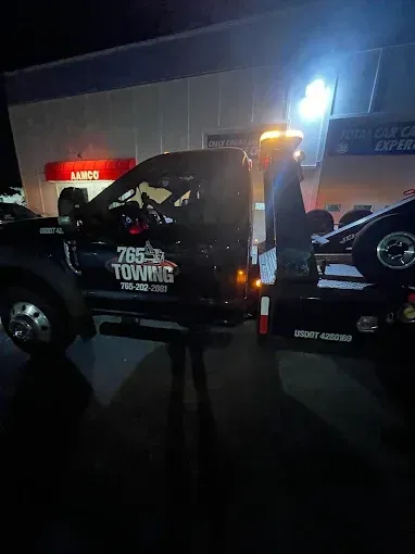Black tow truck with flashing lights towing a car at night in front of a building.