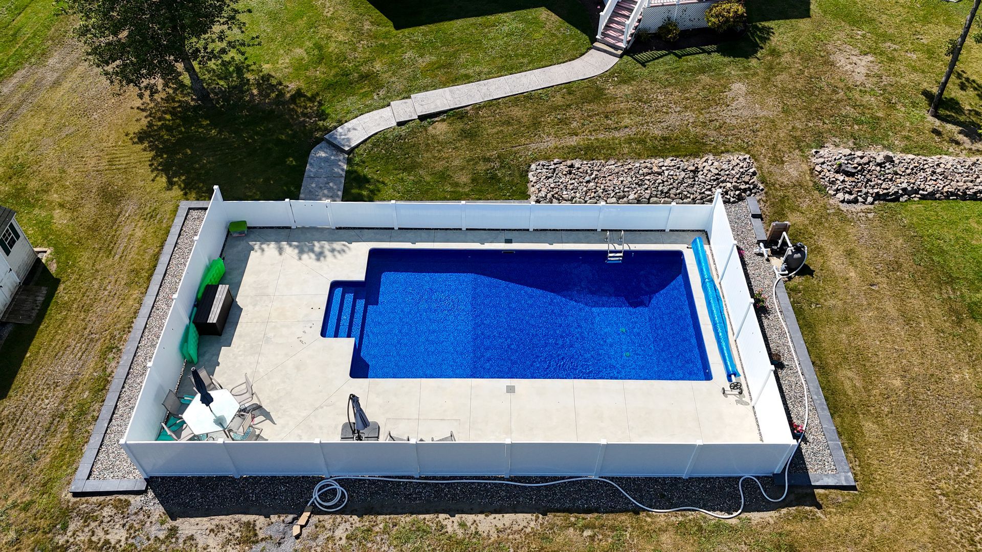 Overhead view of a rectangular pool with blue water, surrounded by a white fence and a concrete patio on grassy land.