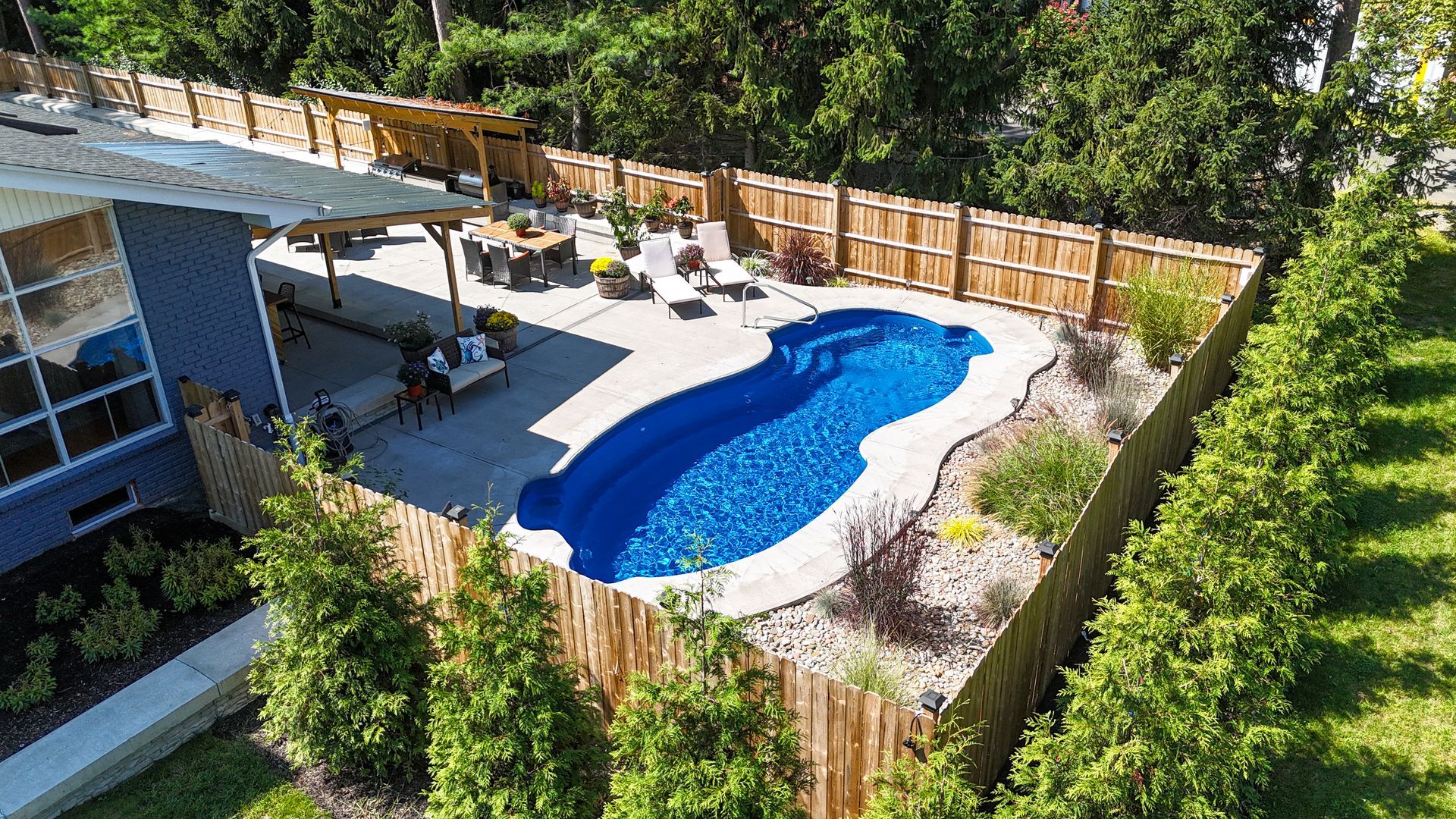 Aerial view of a backyard pool with surrounding wooden fence, patio, and green trees. The pool is filled with blue water.