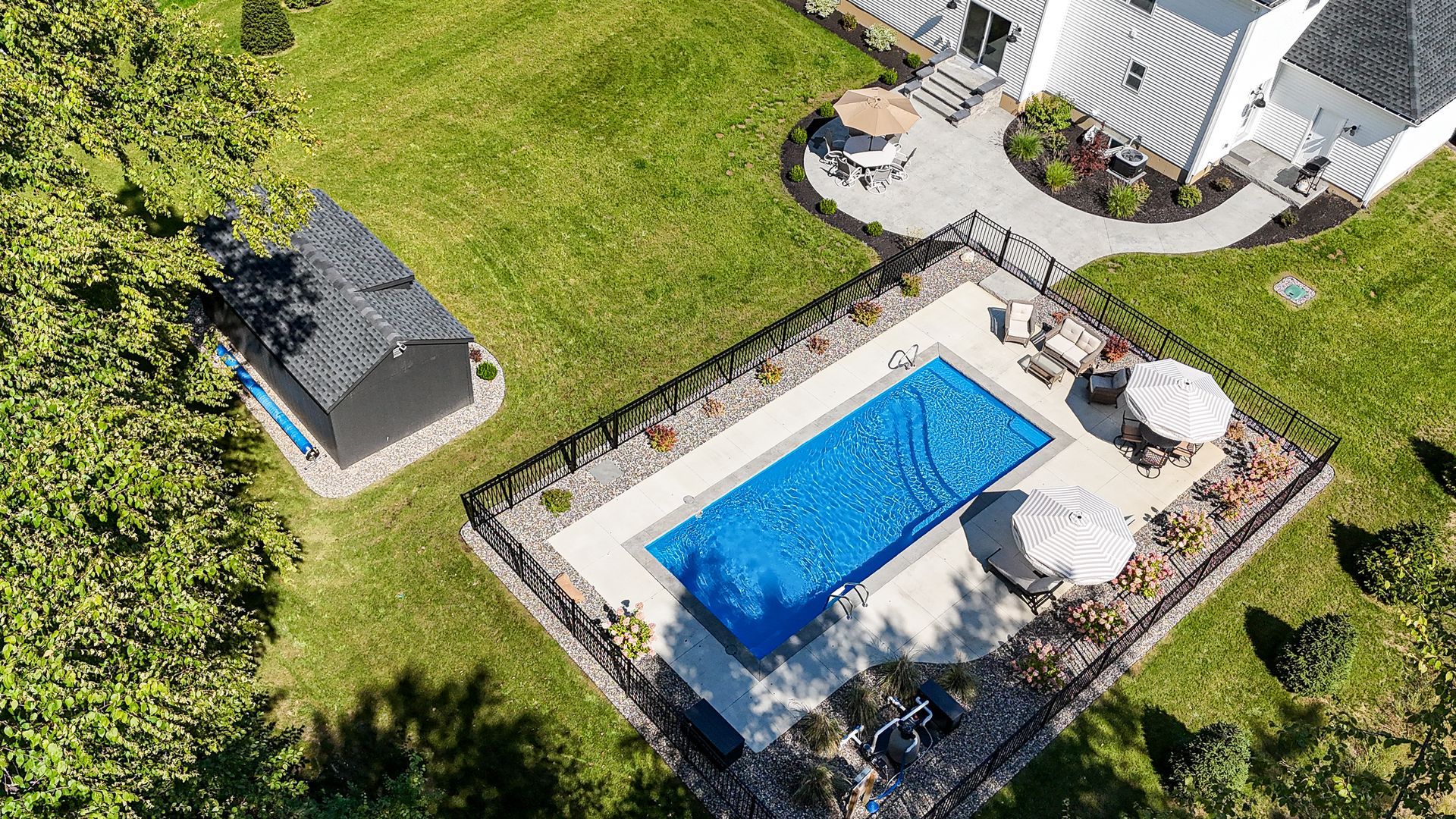 Aerial view: a rectangular swimming pool with blue water and a shed on a green lawn near a white house.
