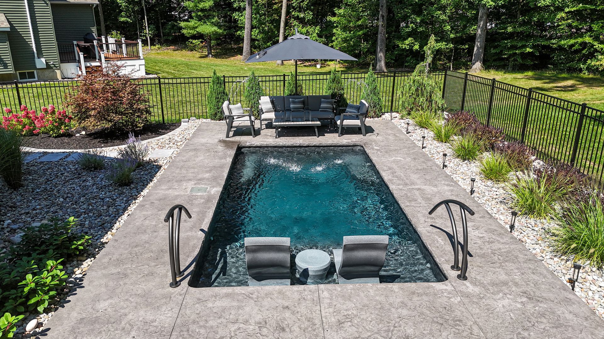 Rectangular pool with seating, a black umbrella, and decorative black fencing in a backyard.