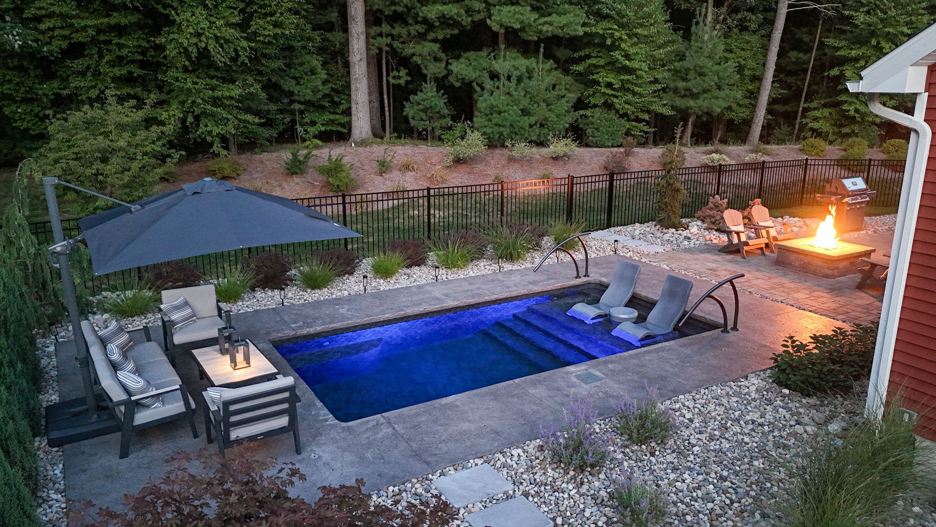 Small rectangular pool lit blue at night, surrounded by patio furniture and a fire pit.