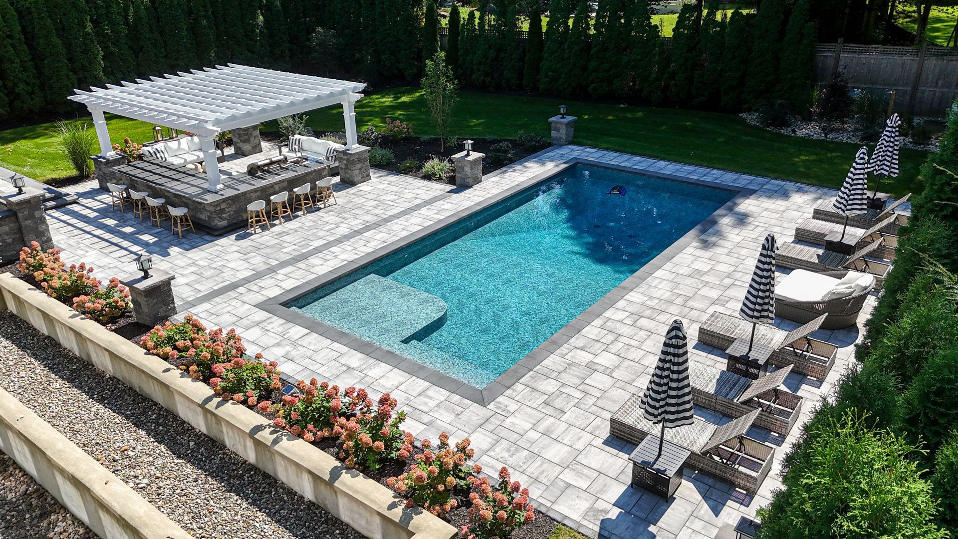 Rectangular pool surrounded by a patio, a covered bar, and lounge chairs in a manicured backyard.