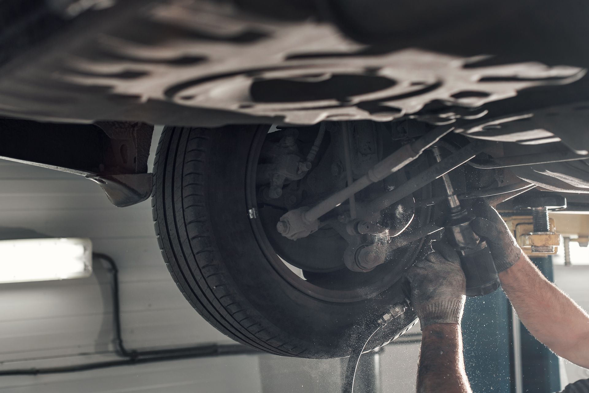 A mechanic in gloves uses a tool to perform maintenance on the lifted wheel and suspension system of a car.