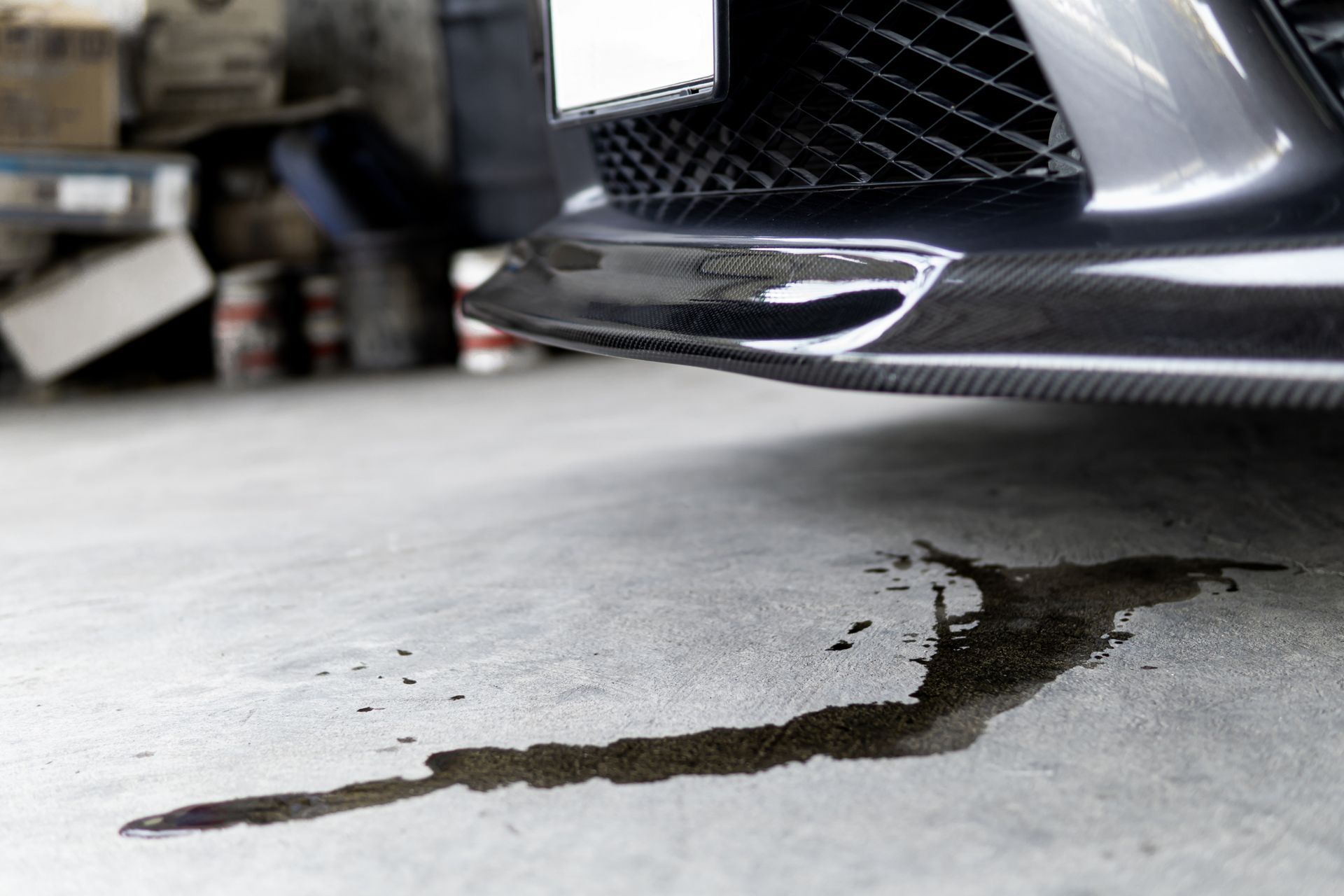 A dark pool of engine oil leaks onto a concrete garage floor beneath the front bumper of a car.